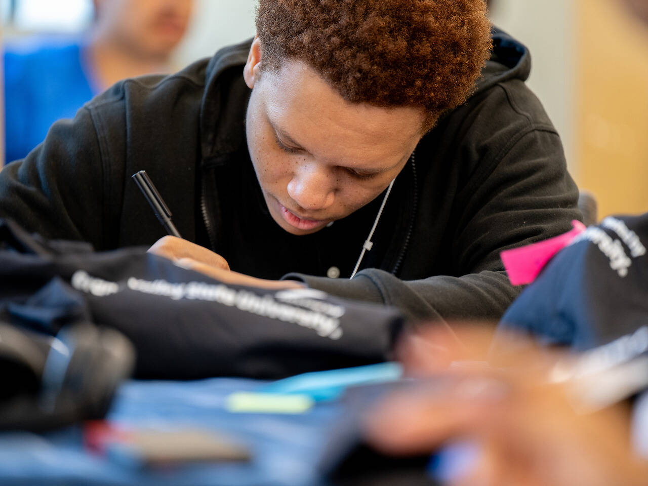 A person with curly hair, wearing a black hoodie, leans over a table signing items while holding a pen.